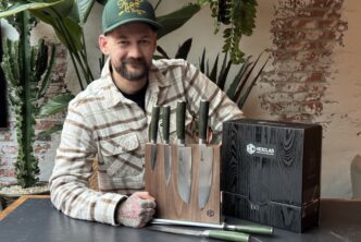Jan Willem Huffmeijer poseert met een HexClad messenblok in een rustieke setting met bakstenen muren en veel groen. Hij draagt een groene pet en een geruite blouse, en leunt ontspannen op de tafel naast de luxe messenverpakking.