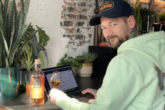 Jan Willem Huffmeijer test de Berry Bros & Rudd 12 Year Old Speyside Sherry Cask whisky terwijl hij aan zijn review werkt op zijn laptop. Hij zit in een sfeervolle setting met veel groen en een industriële bakstenen muur op de achtergrond. In zijn hand houdt hij een whiskyglas met een diepe amberkleurige dram, en naast hem staat de fles prominent op tafel. Hij kijkt met een ontspannen, tevreden blik naar de camera.