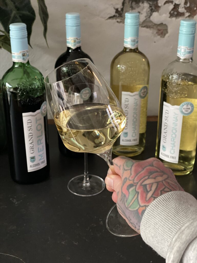 Hand met tattoo houdt een glas Grand Sud alcoholvrije Chardonnay vast
