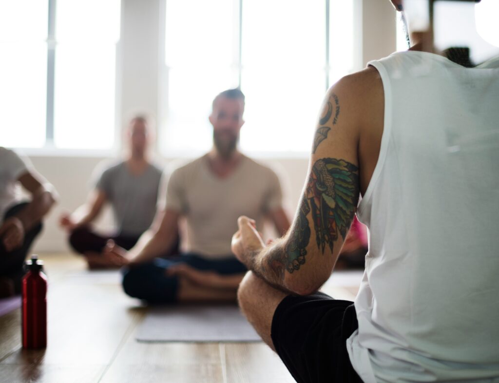 Een groep mannen volgt een Yin Yoga-les, met vooraan een deelnemer met tatoeage die in lotushouding zit. De foto laat zien hoe Yin Yoga rust en concentratie brengt zonder zweverig te zijn.