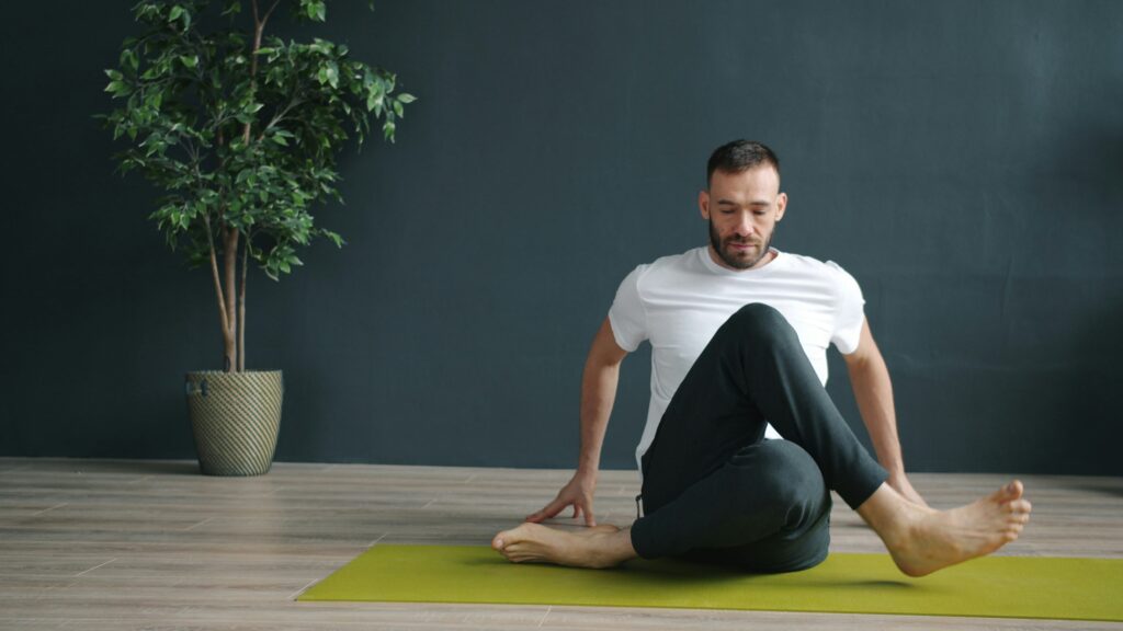 Een man zit op een groene yogamat en voert een Yin Yoga-houding uit in een rustige studio met donkergroene wand en plant ernaast. De foto onderstreept de kalmte en focus van de practice.
